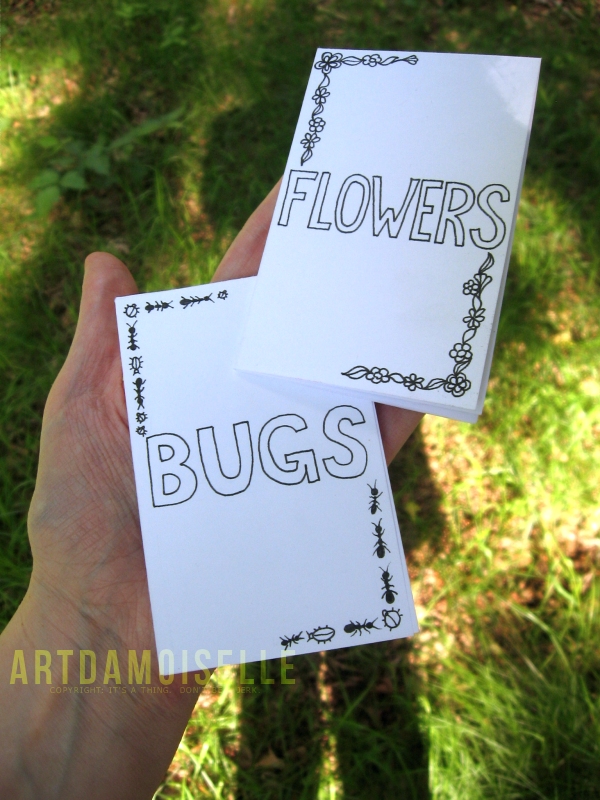 A hand holding two small handmade booklets titled "FLOWERS" and "BUGS".